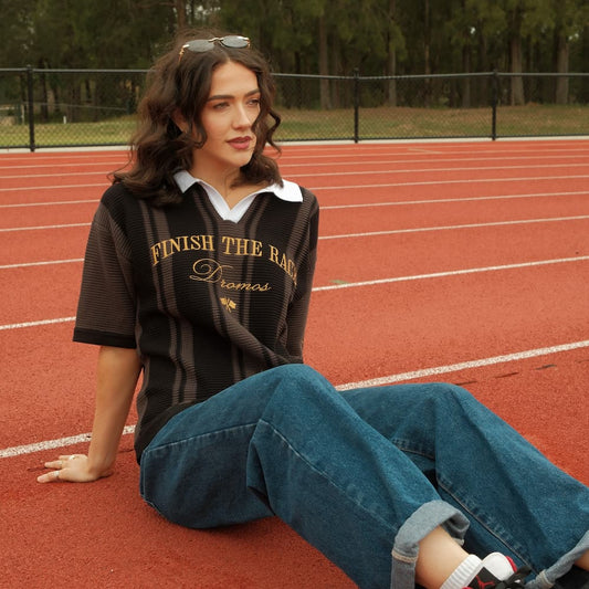Woman sitting on a running track wearing a black christian knit polo with text finish the race and blue jeans.