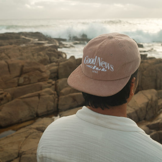 Person wearing a 'Good News Club' cap overlooking rocky cliffs and ocean.