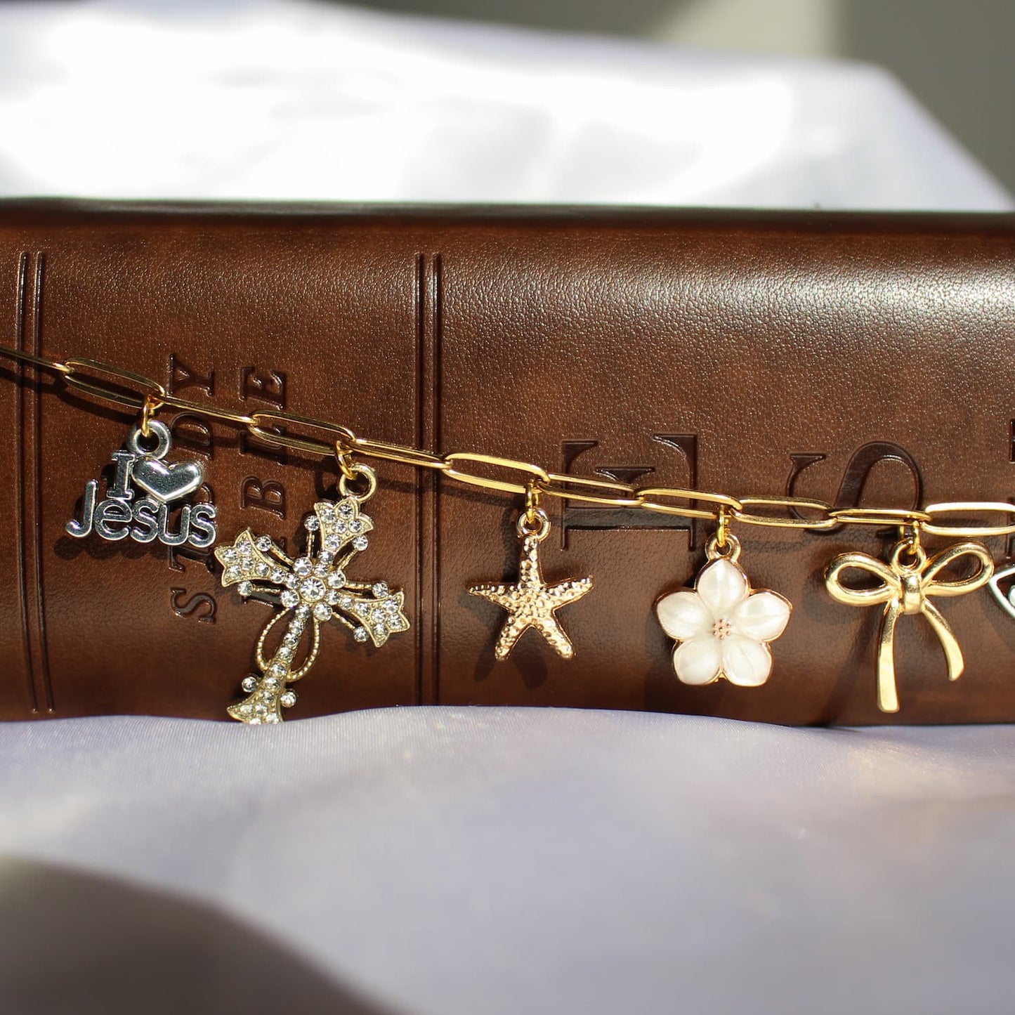 A gold-tone chain bracelet with five dangling charms, including a silver Jesus heart, a silver cross, a starfish, a white flower, and a gold bow, displayed on a brown leather Bible cover.