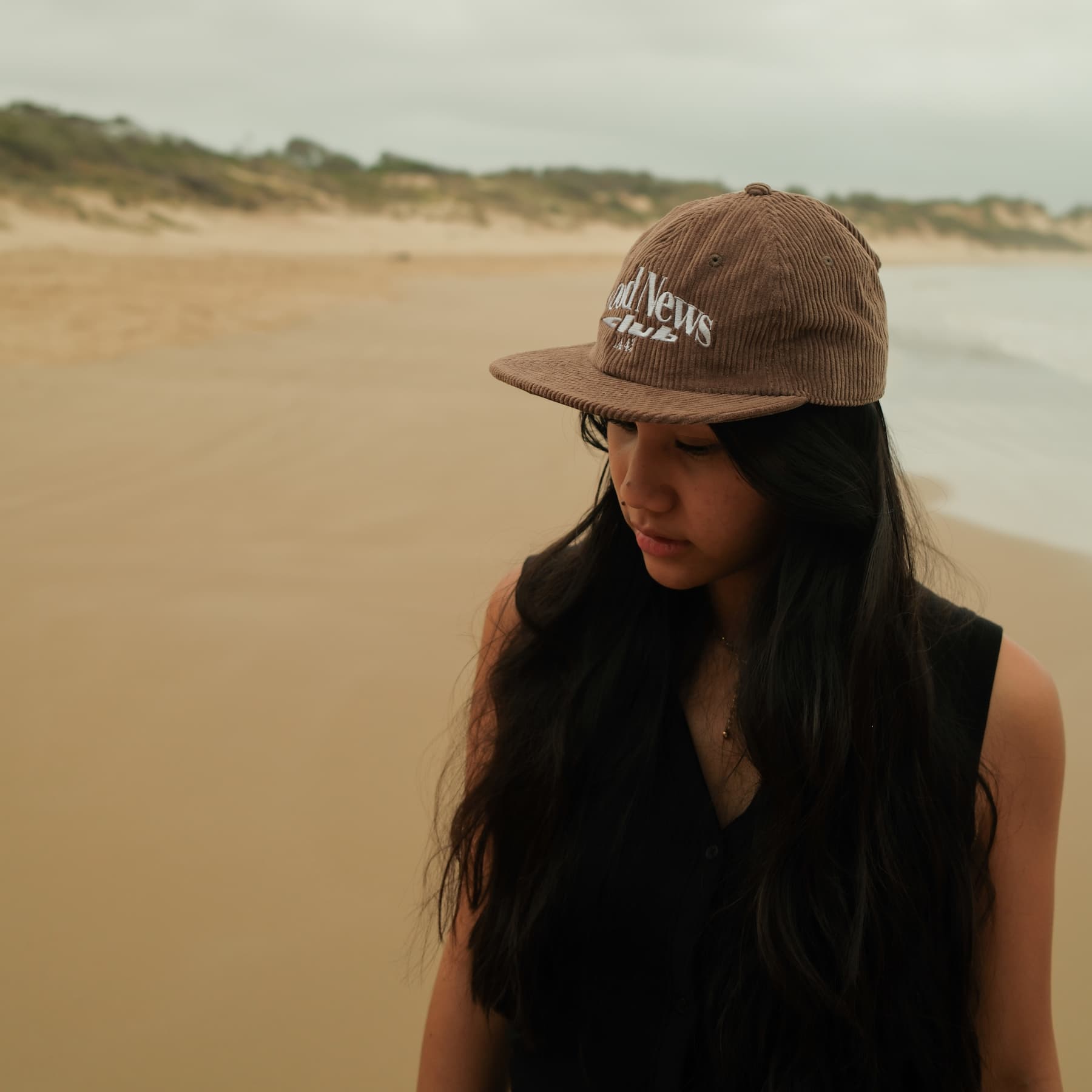 Woman wearing a brown cap with text Good News, standing on a sandy beach.