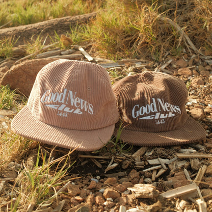 One brown and one hazel pink 'Good News Club' hats on a rustic background with grass and logs.
