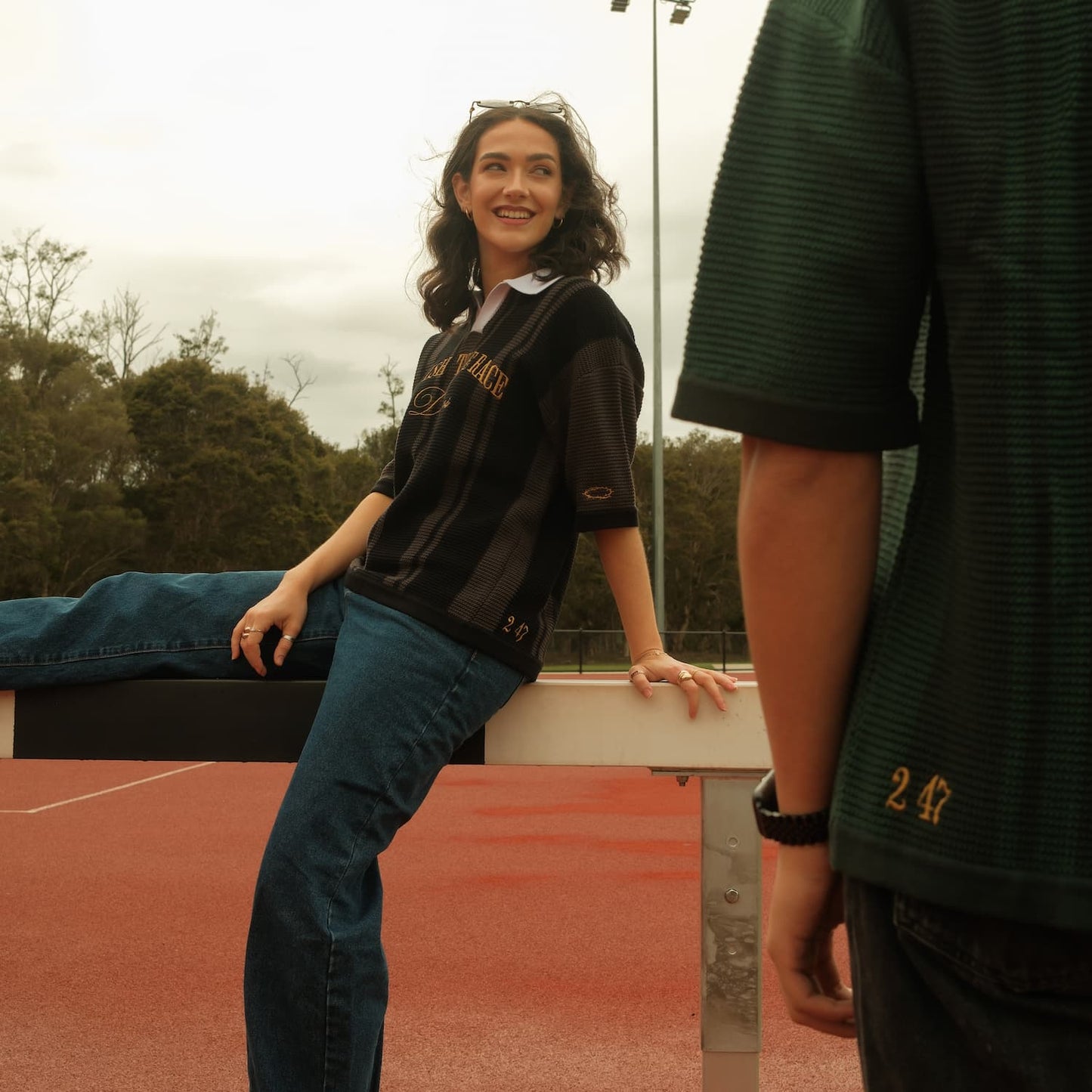 Woman in black knit polo and blue jeans standing on a tennis court with a person in green shirt partially visible.