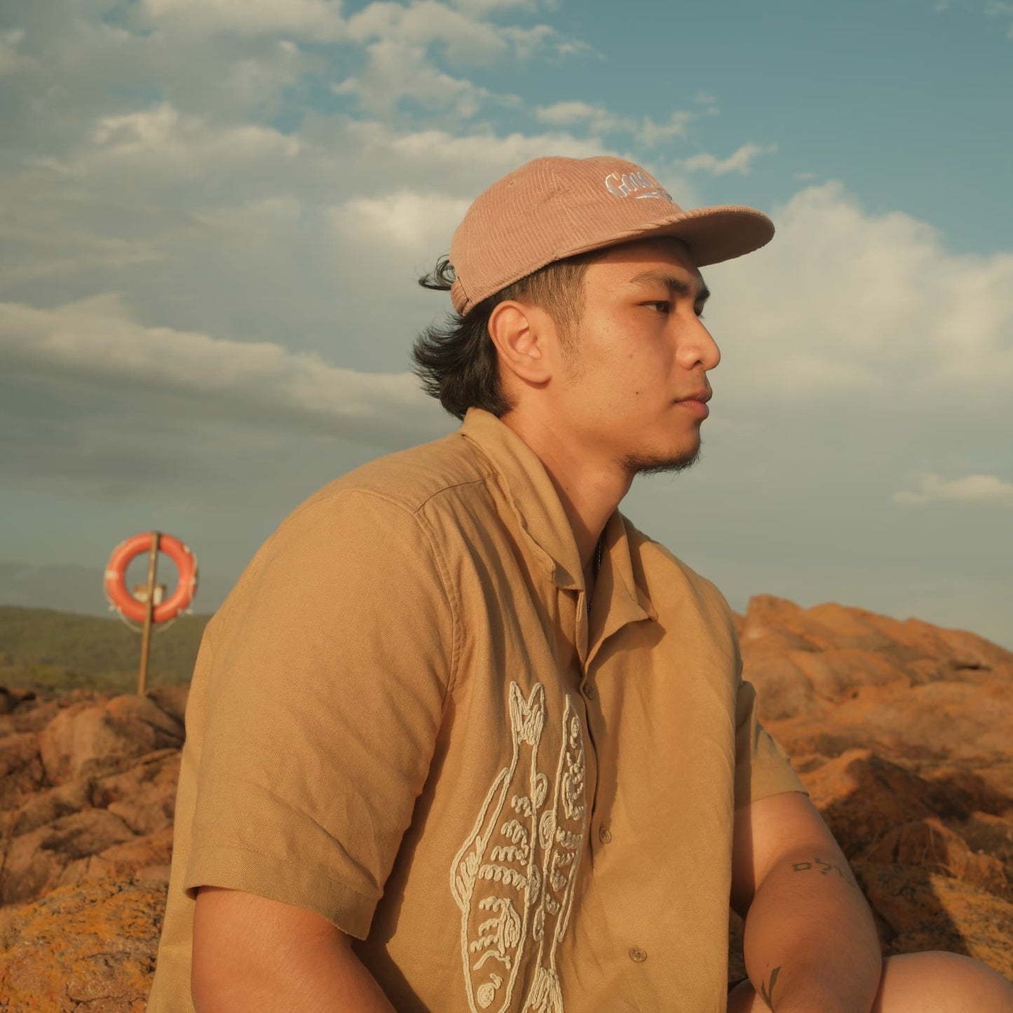 Man wearing a cap and brown shirt sitting on a rocky landscape with a life buoy in the background.