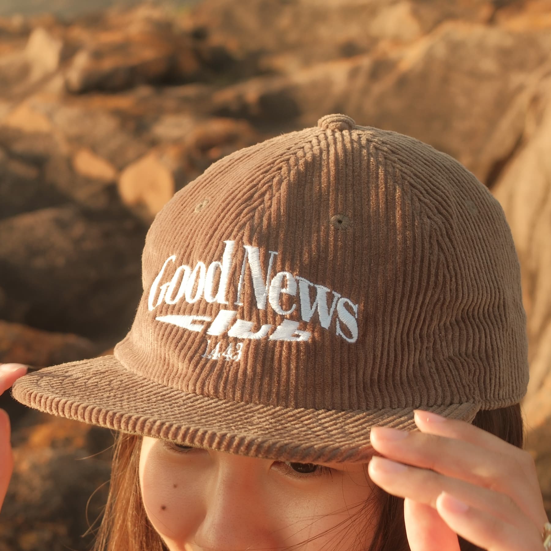 Brown cap with 'Good News Club' text held by a person against a rocky background