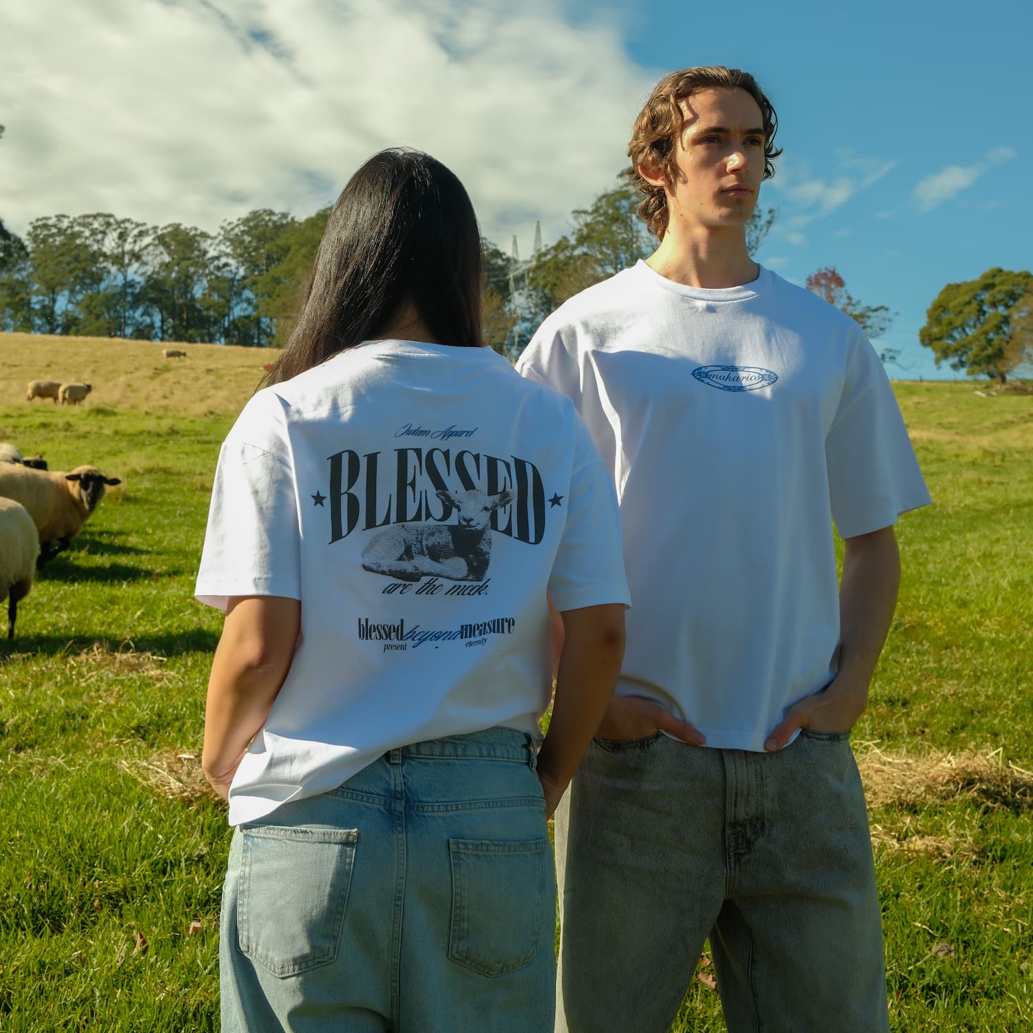 Two people standing in a field with sheep, wearing white t-shirts.