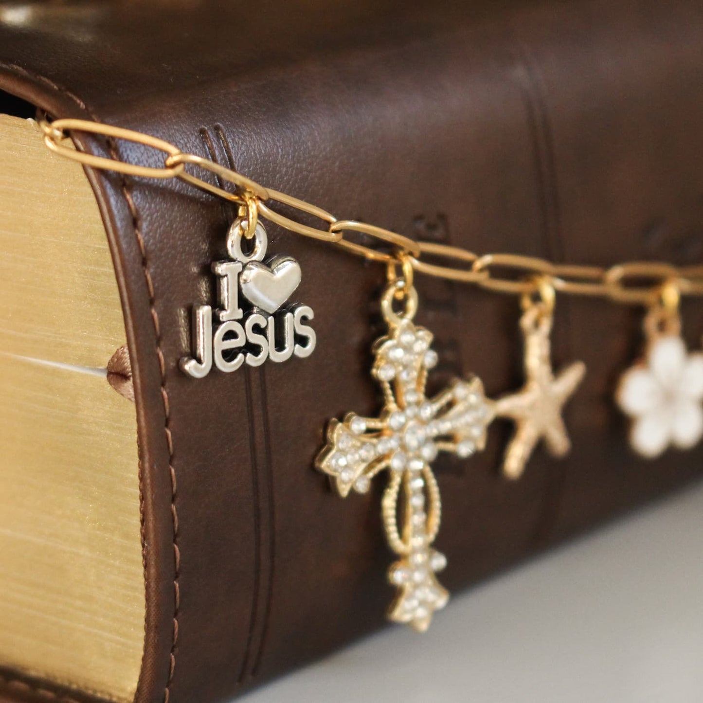 A gold-tone chain bookmark draped over a brown leather-bound Bible, featuring dangling charms including a silver 'I ♥ Jesus' charm, a gold cross with rhinestones, a gold star, and a white flower charm.