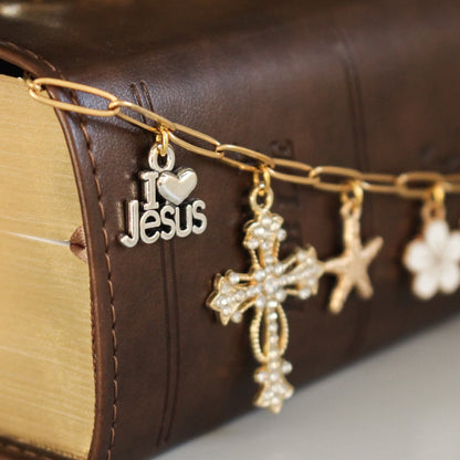 A gold-tone chain bookmark draped over a brown leather-bound Bible, featuring dangling charms including a silver 'I ♥ Jesus' charm, a gold cross with rhinestones, a gold star, and a white flower charm.