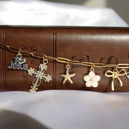 A gold-tone chain bracelet with five dangling charms, including a silver Jesus heart, a silver cross, a starfish, a white flower, and a gold bow, displayed on a brown leather Bible cover.