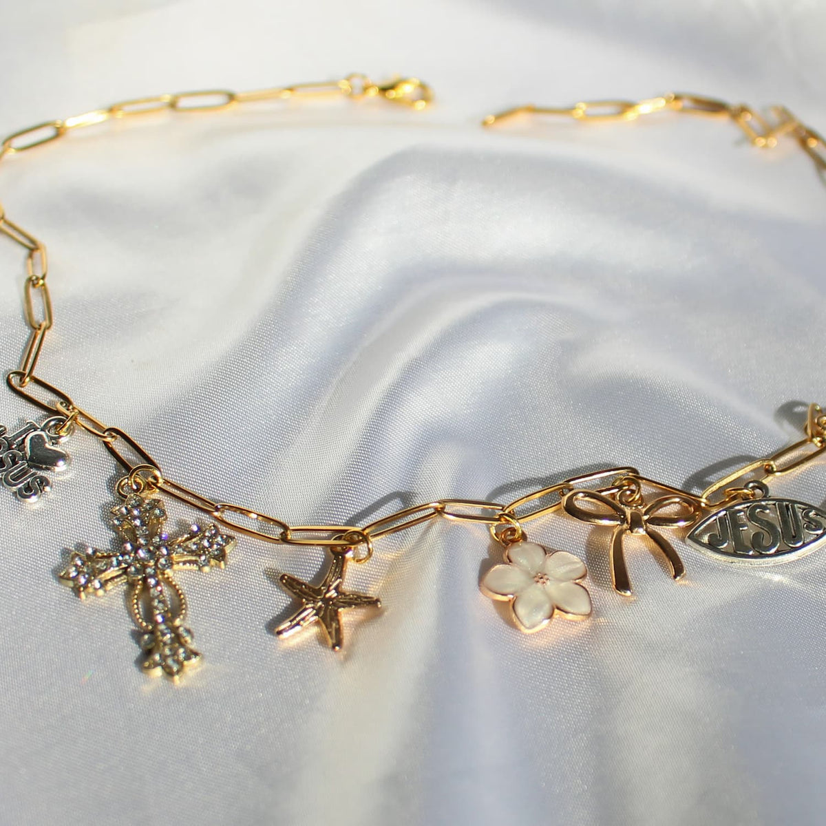 A gold-toned chain bracelet with multiple charms, including a cross, starfish, flower, bow, and fish-shaped pendant, displayed on a white satin fabric background.