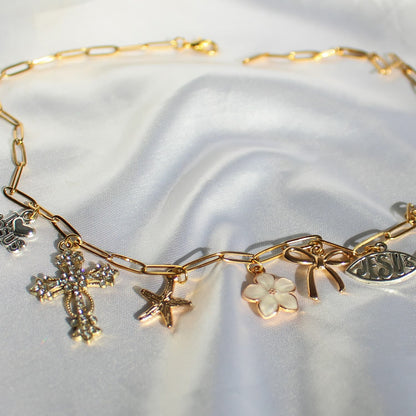 A gold-toned chain bracelet with multiple charms, including a cross, starfish, flower, bow, and fish-shaped pendant, displayed on a white satin fabric background.